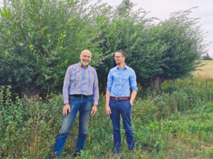 Zwei Männer stehen auf einem Feld vor Kopfweiden mit begrünten Baumkronen
