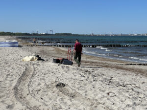 Mann läuft am Strand zu der verteilten Sachen seiner Ausrüstung. Im Hintergrund sind die Buhnen, die Ostsee und die Hansestadt Rostock zu sehen.