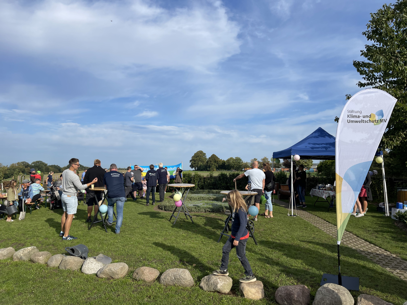 Menschen auf einer Rasenfläche unter freiem Himmel an Stehtischen und auf Bierbänken mit Pavillion und Luftballons