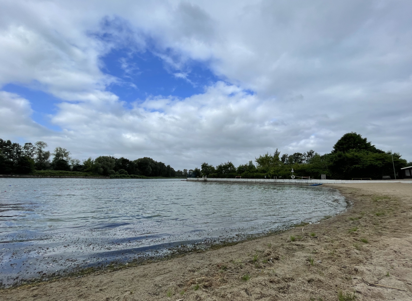 Naturstrand mit Wasser und Bäumen im Hintergrund vor einem Himmel voller Wolken
